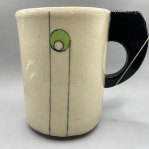 May include: Cream-colored ceramic mug with a black handle and a decorative design. The mug features a green circle with a black outline and two vertical black lines. The handle is black with a white diagonal line.
