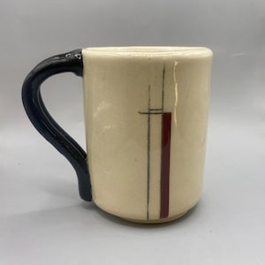 May include: Cream-colored ceramic mug with a black handle. The mug features a vertical design with a red stripe and black lines. The mug is a cylindrical shape.