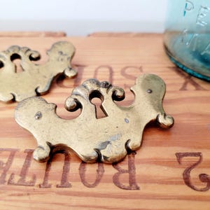 Op de afbeelding: Close-up van drie antieke messing sleutelgatplaatjes. De rozetten hebben een sierlijk, symmetrisch ontwerp met een sleutelgat in het midden. Het messing heeft een warme, verouderde patina. De achtergrond is een houten oppervlak.