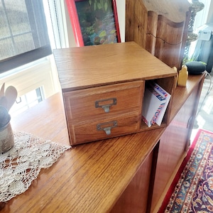 May include: A wooden desk organiser with two drawers and a shelf. The organiser is made of light brown wood and has metal label holders on the drawers. The shelf holds several envelopes. The organiser sits on a wooden surface.