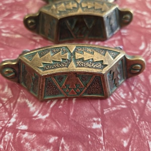 May include: A set of three brass drawer pulls featuring a curved, trapezoidal shape with an aged patina finish. Each pull displays a geometric pattern of embossed arrows and triangles with recessed green accents and two screw holes for mounting.