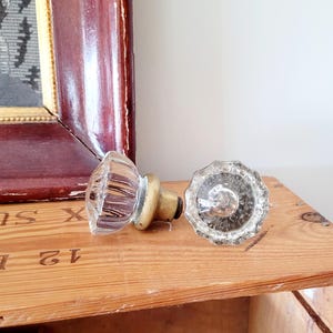 May include: Two vintage clear glass door knobs with brass fittings. One knob is lying on its side, and the other is upright. The knobs are on a wooden surface, with a framed picture in the background.