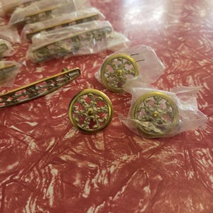 May include: A collection of gold-toned metal cabinet hardware, including several long, curved drawer pulls and circular knobs. Each piece features an intricate, openwork filigree design. Some items are individually wrapped in clear plastic bags.