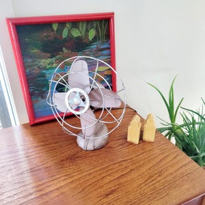 May include: A vintage, light gray electric fan with a silver wire cage and four blades. The fan sits on a wooden surface next to two small, light-colored decorative objects and a framed painting with a red border.