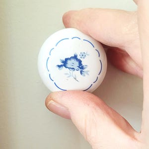 Peut inclure: Un petit bouton rond en céramique blanche avec un motif floral bleu. Le motif présente une fleur bleue avec des feuilles et une bordure bleue festonnée. Le bouton est tenu entre les doigts.