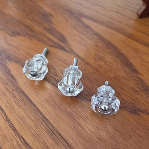 May include: Three clear, faceted glass cabinet knobs with a hexagonal shape and a silver-toned metal screw protruding from the back of each piece. The knobs feature a central silver-coloured screw head visible on the front face of each glass component.