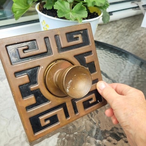 May include: A square brass door plate featuring a central circular knob and a black recessed geometric Greek key pattern. The metal surface shows a weathered patina with visible signs of wear and minor scratches on the knob and frame.