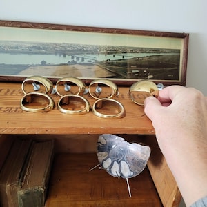 Peut inclure: Ensemble de six poignées de meubles dorées, de forme ovale avec un dessus arrondi, disposées sur une surface en bois. Une main tend la main vers l'une des poignées. Une peinture de paysage encadrée est en arrière-plan.