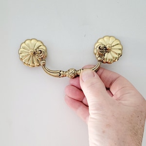 May include: A gold-colored drawer pull with a floral design on each end. The pull has a curved handle and is held by a hand against a white background. The hardware is likely for a cabinet or dresser.