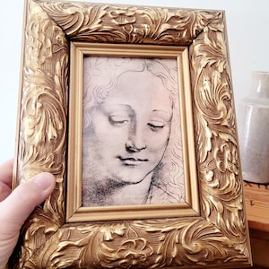 May include: An antique-style gold picture frame with elaborate floral carvings. The frame holds a black and white sketch of a woman's face. The frame is held by a hand.