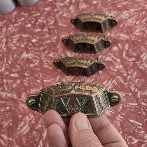 May include: Four brass drawer pulls featuring a geometric pattern with triangular motifs and a dark patina finish. Each pull has a curved, elongated shape with two screw holes on the ends for mounting, showcasing a textured, aged metallic surface.