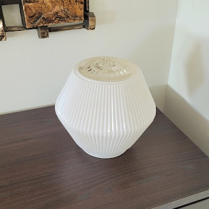Peut inclure: Une lampe en verre blanc côtelé avec un dessus décoratif est posée sur une surface en bois brun foncé. La lampe a une forme géométrique et un dessus détaillé à motifs. Un miroir décoratif est en arrière-plan.