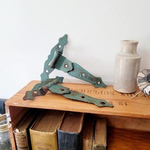 Può includere: Due antiche cerniere in metallo color turchese con bordi decorativi e ritagli a forma di diamante. Le cerniere sono posizionate su una cassa di legno con vecchi libri visibili sotto. Sono presenti anche un vaso di ceramica e un oggetto decorativo.