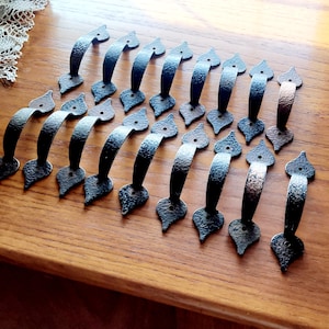 May include: A collection of black metal handles with a hammered texture. Each handle features a curved grip and decorative end pieces. The handles are arranged on a wooden surface, showcasing their design and potential use in furniture or cabinetry.