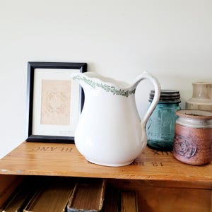 May include: White ceramic pitcher with a scalloped rim and green floral detailing. The pitcher has a curved handle and sits on a wooden shelf with other vintage items, including a framed print and a blue glass jar.