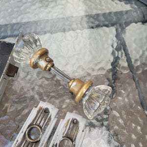 May include: A vintage door hardware set featuring two clear, fluted glass knobs with brass-toned metal bases connected by a central spindle, accompanied by two rectangular white metal backplates with circular openings and visible screw holes.
