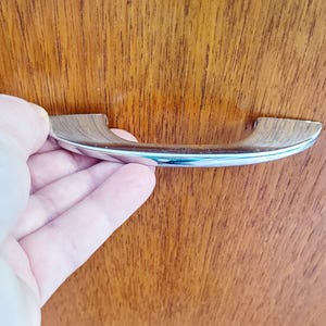 Peut inclure: Une poignée de meuble chromée et aspect bois. La poignée est tenue contre une surface en bois. La poignée a un design incurvé avec une finition chromée à l'avant et une finition aspect bois à l'arrière. La surface en bois a un grain naturel.