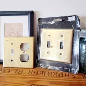 May include: Two vintage brass electrical switch plates. One plate has an outlet and a switch, the other has two switches and is still in its original packaging. The plates are on a wooden surface.
