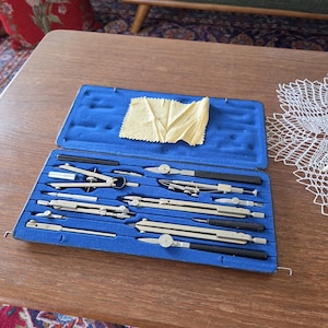 May include: A vintage drafting instrument set housed in a rectangular case with a blue velvet interior. The kit includes various metal compasses, dividers, and extension bars, along with a small yellow cleaning cloth tucked inside the lid.