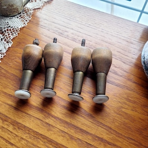 May include: Four vintage wooden furniture legs with brass-colored metal shafts and white plastic feet. The legs have a turned, bulbous shape and are designed for easy attachment to furniture. The wood has a warm, natural finish.
