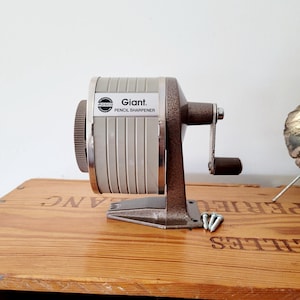 May include: A vintage pencil sharpener with the text "Giant. PENCIL SHARPENER" on a light gray and silver body. The sharpener has a brown crank and base, and is mounted on a wooden surface. Includes three silver screws.