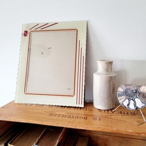 May include: An antique cream-colored picture frame with a scalloped edge and red accents. A small, beige ceramic vase and a decorative, shell-shaped object with metal legs sit nearby. The frame is empty, ready for a photo.