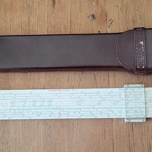 May include: A vintage slide rule and a brown leather case. The slide rule is white with black markings and a clear plastic cursor. The case is rectangular and has a strap with a buckle. The items are on a wooden surface.