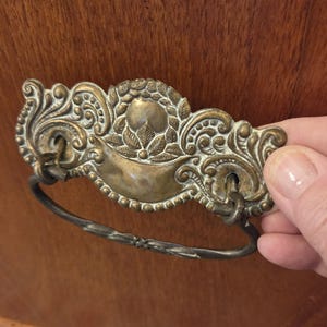 May include: A brass drawer pull featuring an ornate, embossed backplate with intricate scrollwork and leaf motifs. The hardware includes a swinging, oval-shaped drop handle attached to the backplate, showcasing a weathered, aged metal finish.