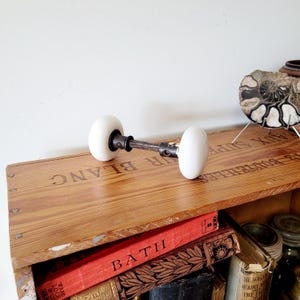 May include: A vintage door handle set featuring two smooth, round white porcelain knobs connected by a dark, weathered metal spindle. The hardware exhibits signs of age and use, with a simple, functional design intended for a standard door mechanism.