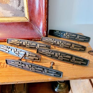 以下が含まれることがあります： 幾何学模様のブロンズトーンのキャビネットプルコレクション。プルは長方形で、一連の隆起した線形形状があります。ハードウェアは木製の表面に配置されており、ヴィンテージまたはレトロな美学を示唆しています。