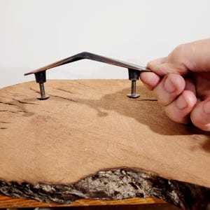 May include: A dark bronze-toned metal drawer pull with an angular, peaked centre design. The handle features two cylindrical mounting posts extending downward, each equipped with a threaded screw and a flat circular nut at the base for attachment.