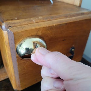 以下が含まれることがあります： 光沢のある金色のノブが付いた木製の箱のクローズアップ。箱は明るい茶色の木材でできており、シンプルなデザインです。ノブは丸く、周囲の環境を反射しています。箱はより大きな家具の一部であるようです。