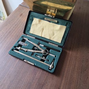 May include: A black Techmatic drafting compass set in a rectangular case with a dark green velvet interior. The set includes silver metal compasses, a small container of lead refills, and a light yellow cleaning cloth attached to the lid.