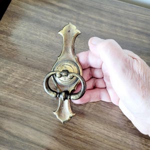 May include: An antique brass door knocker featuring an elongated, decorative backplate with a pointed top and bottom. A circular central mount holds a hinged, oval-shaped ring that hangs downward, showcasing a weathered, dark metallic patina finish.