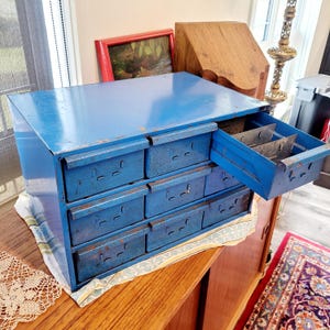 May include: A vintage blue metal cabinet with multiple drawers, one of which is partially open. The cabinet sits on a wooden surface, showcasing its aged, distressed appearance. The cabinet's color is a vibrant blue, with visible signs of wear.