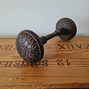 Pode incluir: Um puxador de porta antigo, castanho escuro, com um design circular ornamentado. O puxador é feito de metal e tem uma superfície detalhada e texturizada. O puxador da porta está sobre uma superfície de madeira.