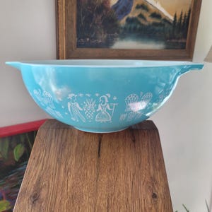 以下が含まれることがあります： 白の人物とバスケットの様式化されたデザインが施された、ターコイズ色のパイレックスミキシングボウル。ボウルは広いリムと両側にハンドルが付いています。このヴィンテージキッチン用品は、混ぜたり、盛り付けたりするのに最適です。