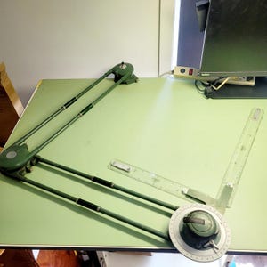 May include: A vintage green drafting table with a parallel rule and protractor. The table has a light green surface and a metal frame. The parallel rule is clear with black markings. The protractor is white with black markings.