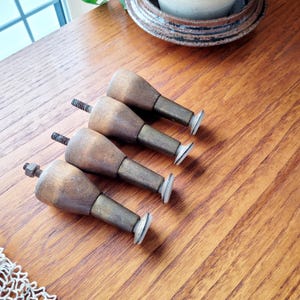 May include: Four vintage wooden furniture legs with brass-colored metal shafts and small, white, circular feet. The legs have a turned, rounded design and are attached with threaded bolts and nuts. The legs are arranged diagonally on a wooden surface.