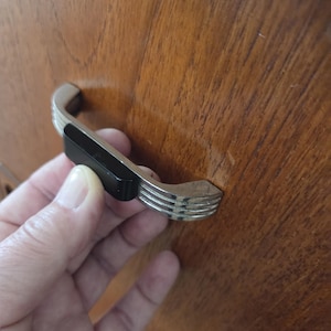 May include: A drawer pull handle featuring a polished silver-tone metal finish with three horizontal ribbed lines on each end and a solid black rectangular center section, mounted on a smooth, medium-brown wood grain surface.