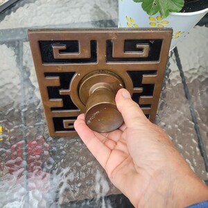 May include: A square brass door plate featuring a geometric Greek key pattern with black inlay and a central circular brass knob. The metal surface shows signs of age and wear with a tarnished patina throughout the hardware components.