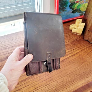 May include: A dark brown leather satchel with a flap closure and a buckle strap. The satchel has a rectangular shape and appears to be vintage. The leather shows signs of wear, adding to its character. The satchel is held by a person.