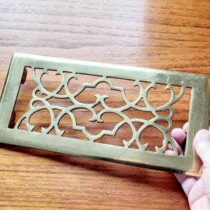 May include: A rectangular brass vent cover with an ornate, openwork design. The cover features a repeating pattern of swirling shapes and curves. The brass has a warm, golden hue, and the edges are slightly rounded.