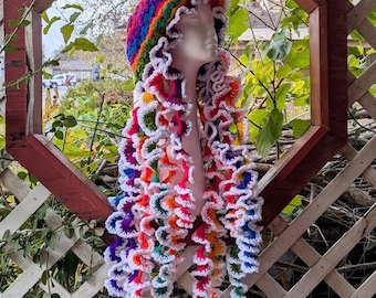 May include: A colorful crocheted hat with a long, flowing scarf attached. The hat and scarf are made with a variety of bright colors, including pink, purple, blue, green, and yellow. The hat is sitting on a white wooden lattice.