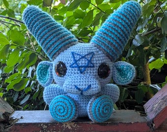 May include: A light blue crochet goat doll with large blue horns, a pentagram, and spiral feet. The handmade plushie is sitting on a wooden ledge with a white lattice fence and green foliage in the background.