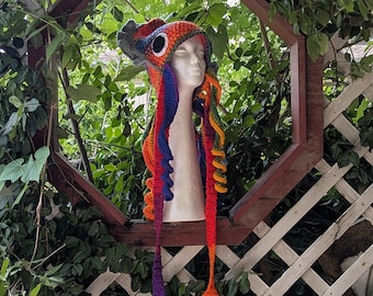 May include: A colorful, handmade crochet hat with a dragon-like design. The hat features a gray headpiece with a large eye, and long, flowing strands in shades of red, orange, yellow, blue, and purple. The hat is displayed on a mannequin head.