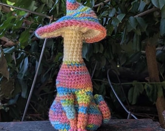 May include: A crocheted figurine featuring a cat-like body with a long neck and a wide-brimmed hat. The body and hat are crafted from yarn in a variegated pattern of pink, blue, yellow, and orange, while the neck is a solid cream color.