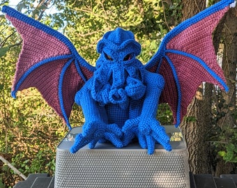May include: A crocheted blue and purple Cthulhu-inspired creature with large wings. The creature is sitting on a gray speaker.