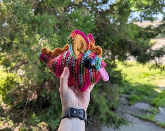 May include: A colorful, handmade crochet creature with a pink tongue, large black eyes, and multicolored yarn. The creature has orange, pink, and blue accents. The item is held in a hand against a blurred green background.