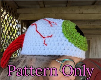 May include: A crochet pattern for a white eye hat with a green eye and red veins. The hat has a red crocheted brim. The pattern is for sale, and the text "Pattern Only" is visible in the image.